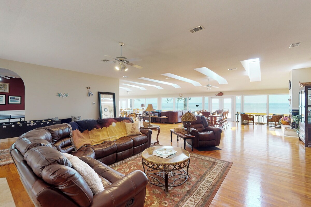 A spacious living room with a large brown leather sofa, a round coffee table, and a view of the ocean through large windows.