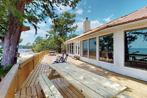 Una terraza de madera con bancos y sillas, con vistas a un cuerpo de agua y una playa.