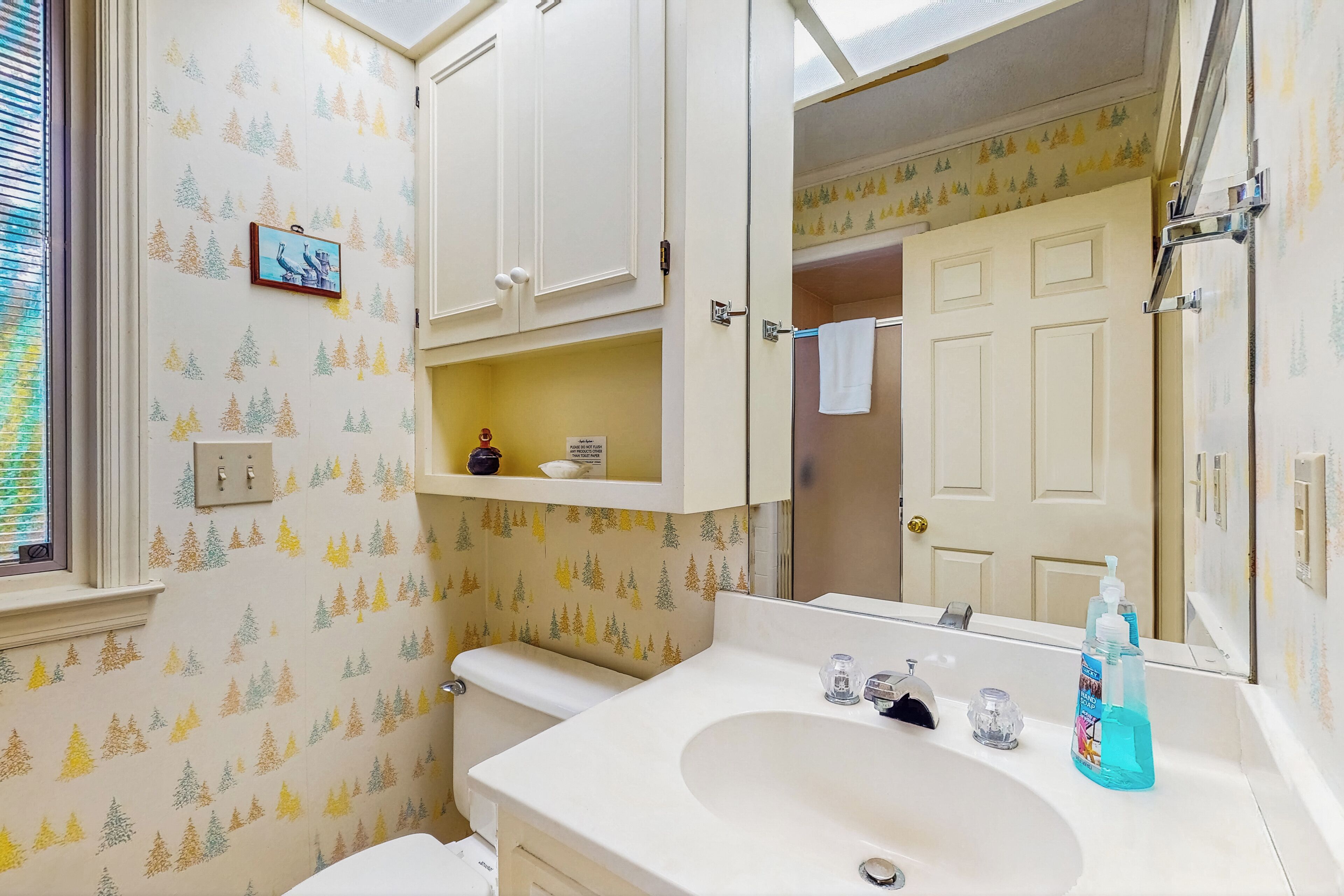 A bathroom with a toilet, sink, and mirror. There is a shelf above the sink and a picture on the wall.