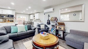 Een moderne woonkamer met een grijze bank, een ronde houten salontafel en een grijze fauteuil. Er is een keuken met witte kasten en een magnetron. Een gitaar hangt aan de muur, en een boekenplank met boeken en een gitaarkoffer is zichtbaar.