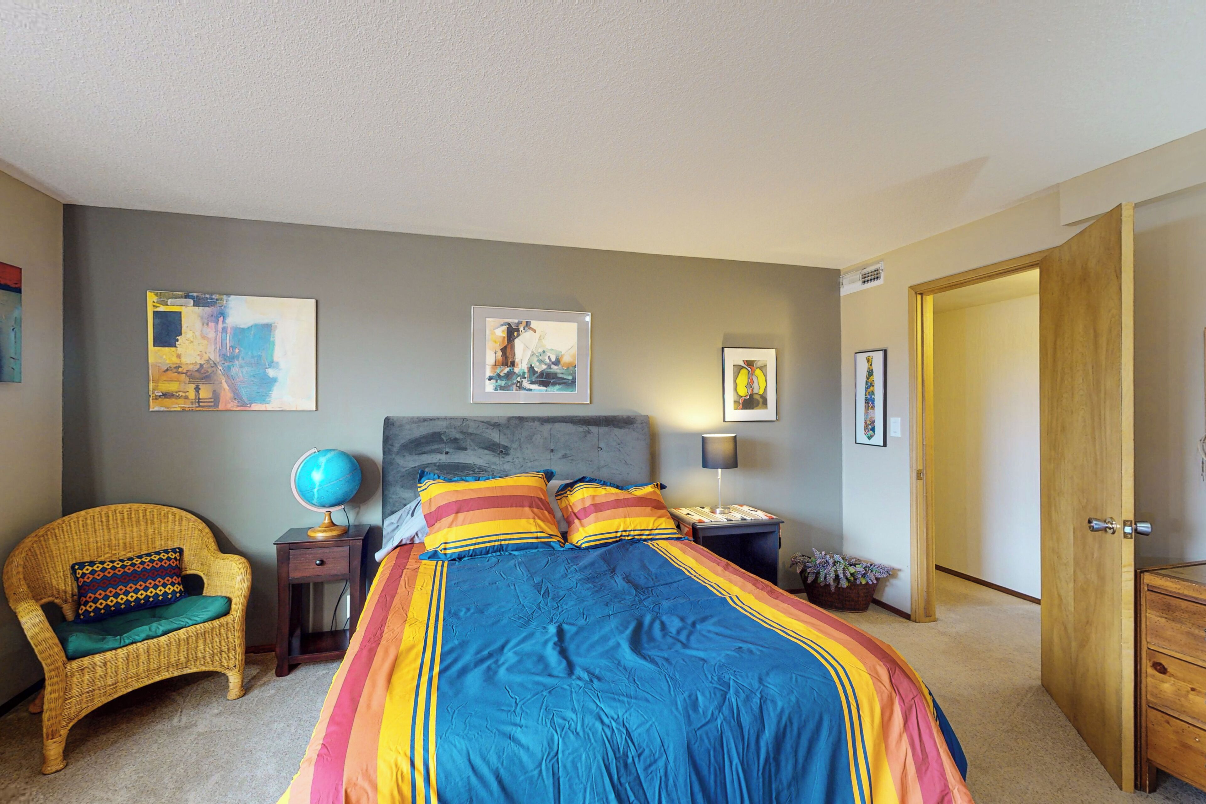 A bedroom with a large bed, a wicker chair, a globe, and artwork on the walls.