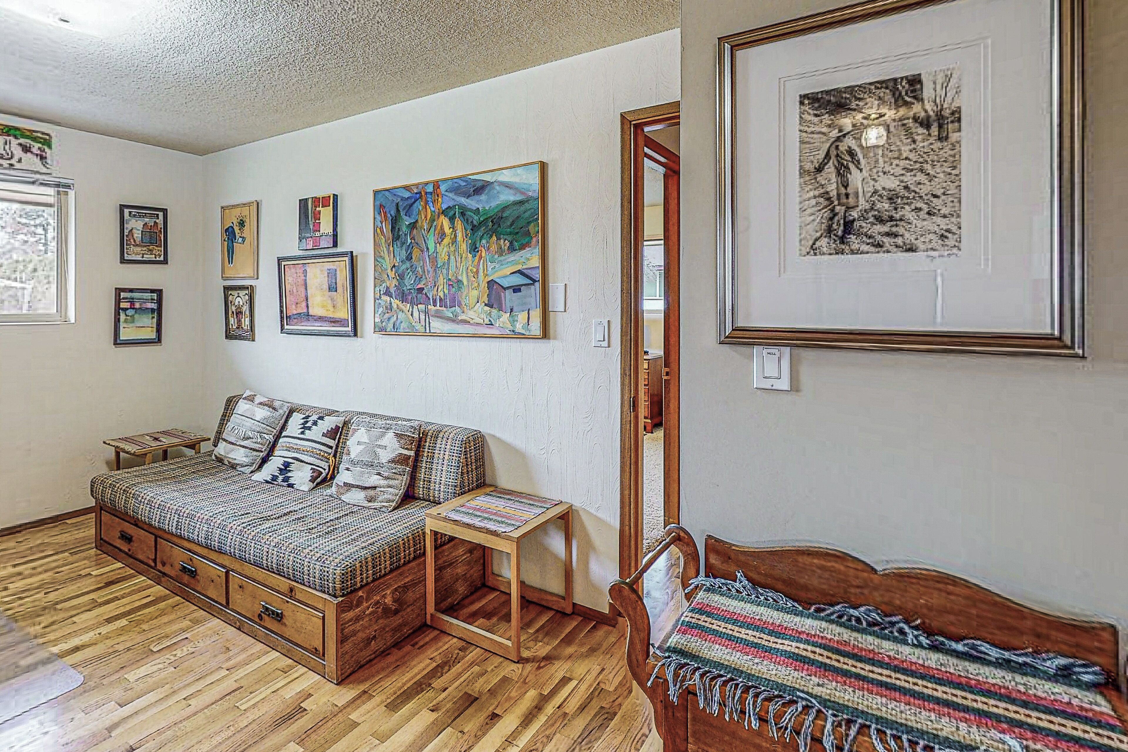 A cozy room with a wooden sofa, a small table, and a bed with a colorful blanket. There are several framed artworks on the walls.