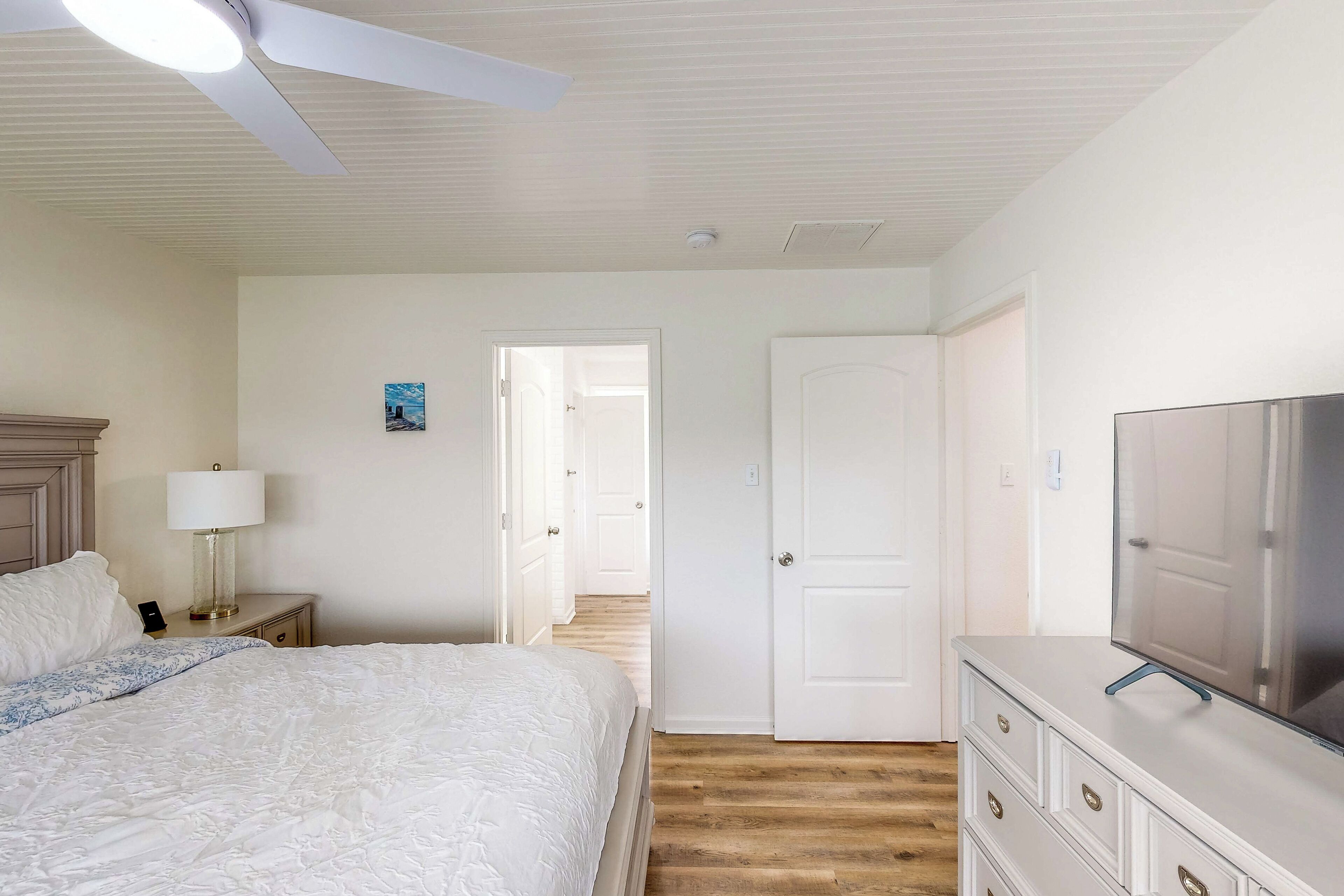 A bedroom with a bed, a nightstand with a lamp, a dresser with a TV, and a door leading to another room.