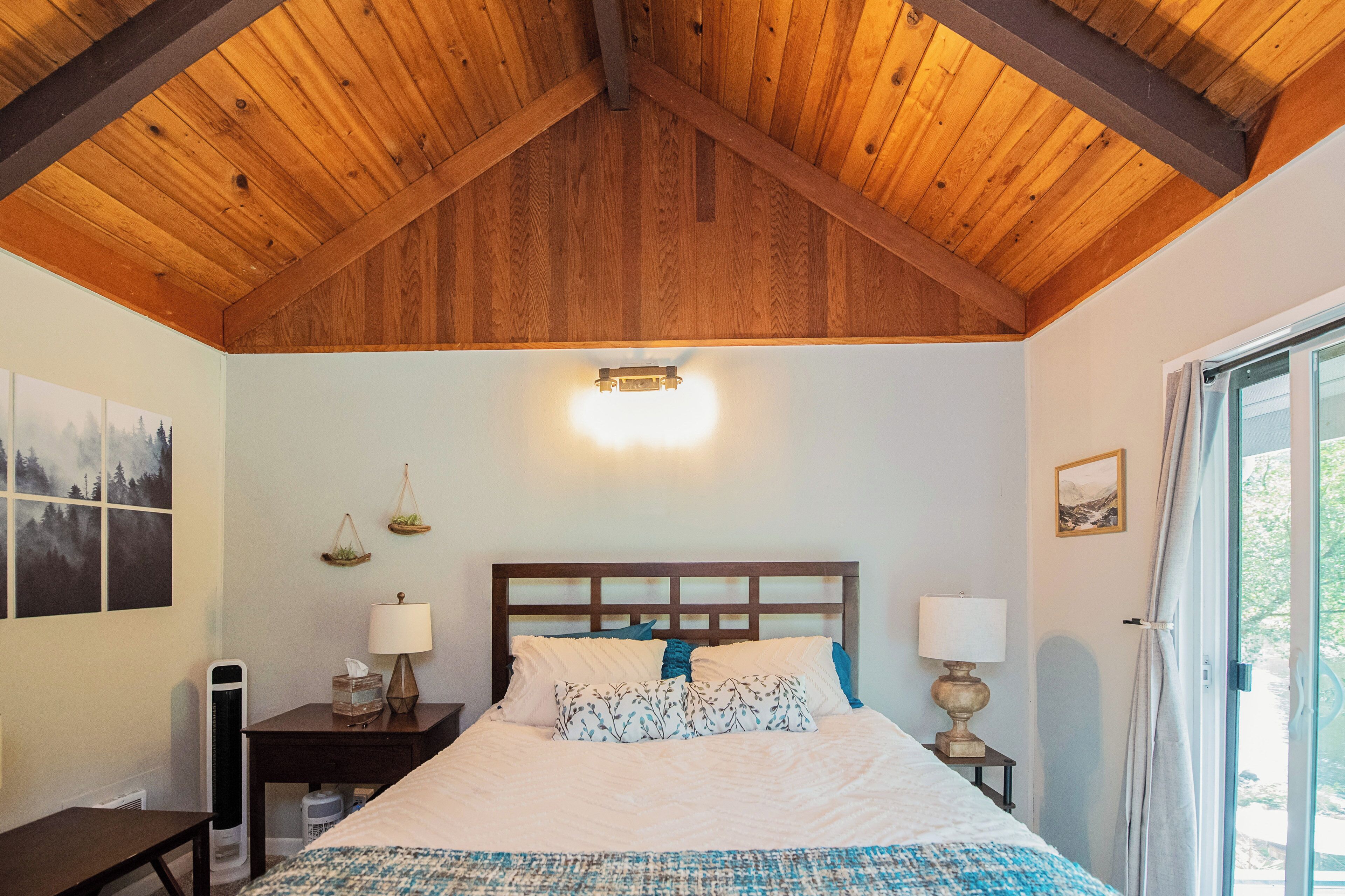 Un dormitorio con techo de madera, una cama con ropa de cama blanca, dos mesitas de noche con lámparas y una ventana con cortinas.