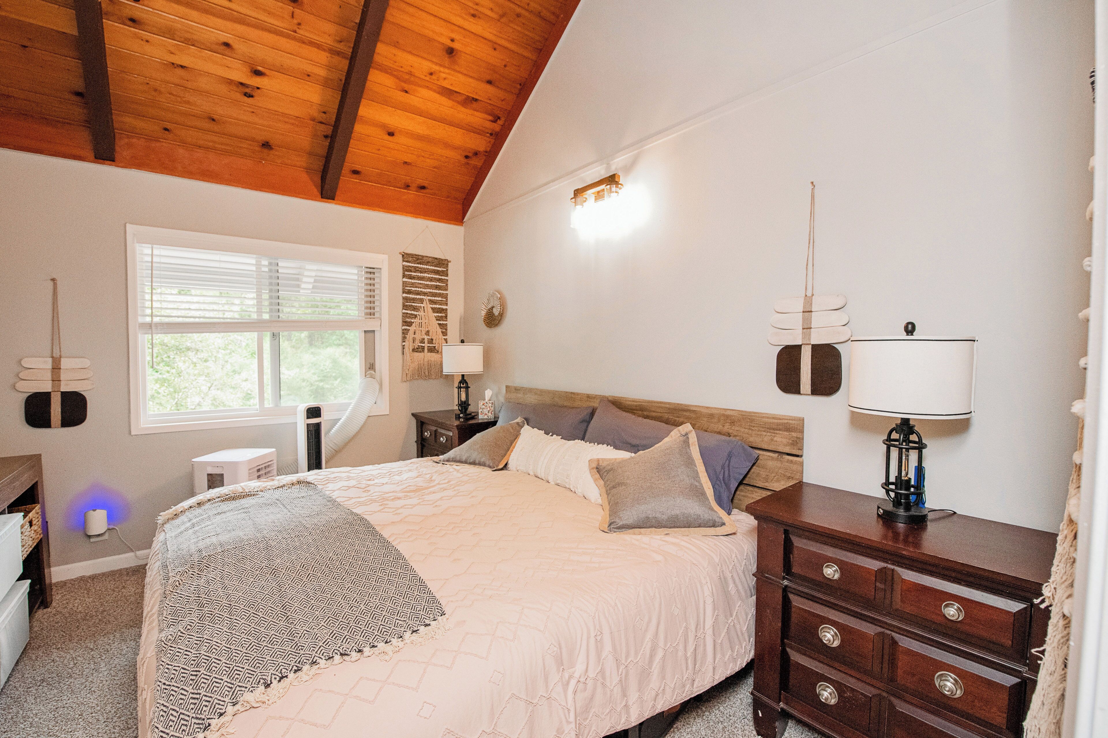 A bedroom with a wooden ceiling, a large bed with patterned bedding, a nightstand with a lamp, and a dresser.