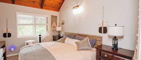A bedroom with a wooden ceiling, a large bed with patterned bedding, a nightstand with a lamp, and a dresser.