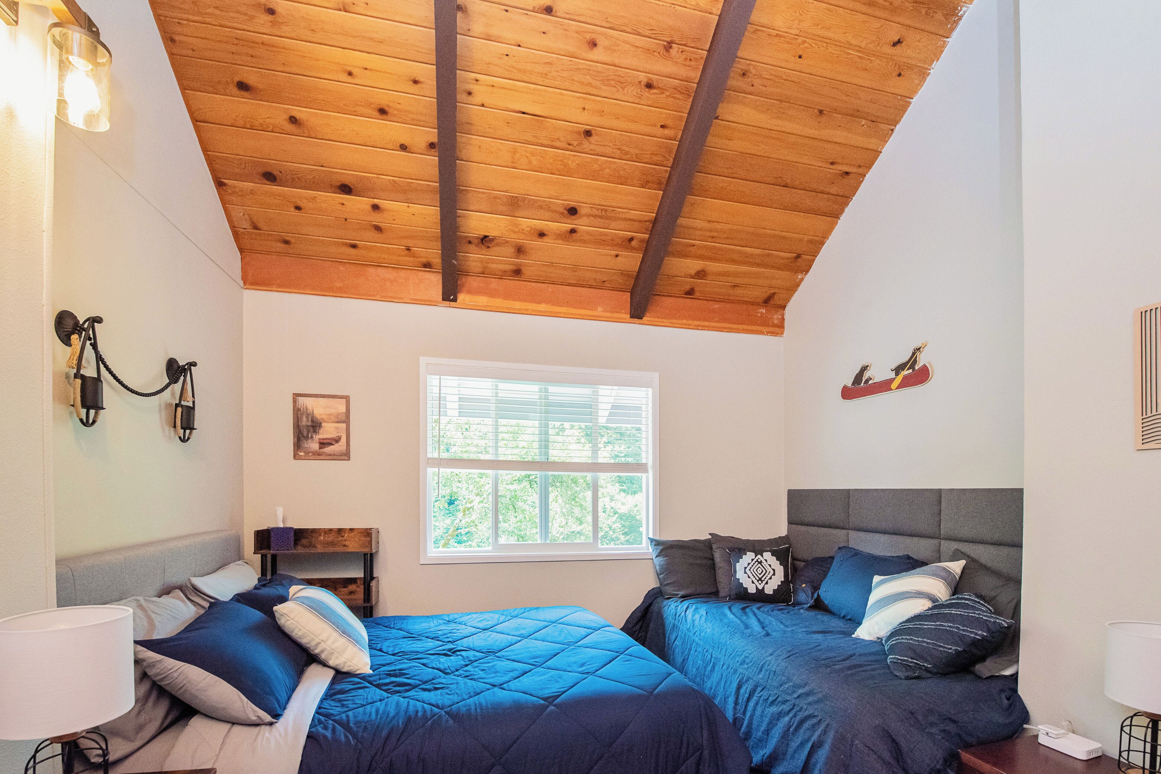 A bedroom with two beds, wooden ceiling beams, a window, and wall-mounted lights.