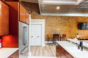 Una cocina con armarios de madera, un refrigerador de acero inoxidable, una puerta blanca, una pared de ladrillo, una mesa pequeña con sillas y un televisor montado.