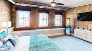 A bedroom with exposed brick walls, a large bed with a blue and white bedspread, a ceiling fan, and a TV mounted on the wall. - 'downtown Delight' w/ Patio! 1 Mi to Ball State (Muncie)
