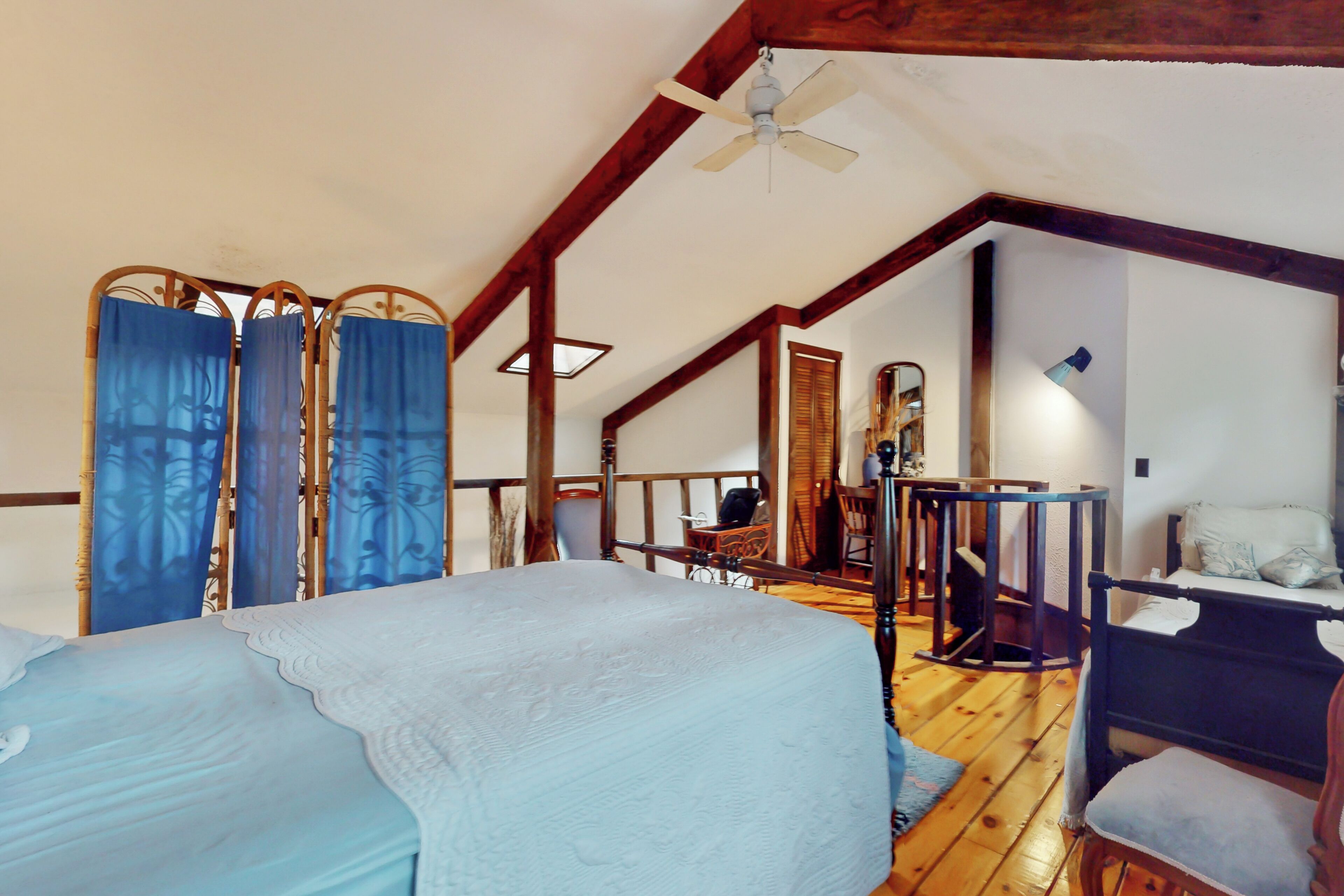 A bedroom with a bed, a desk, and a chair. There is a ceiling fan and a window with curtains.