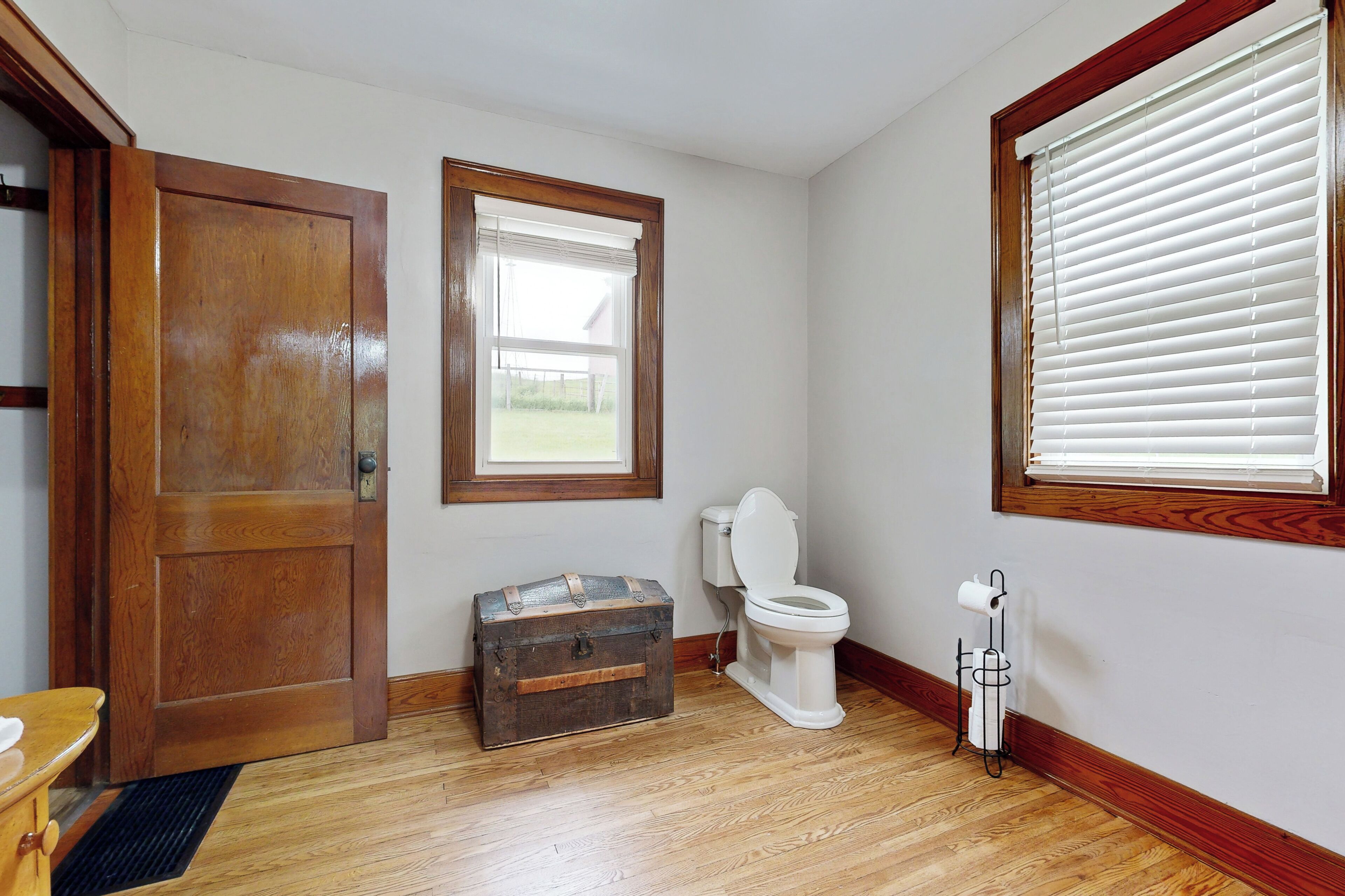 Une salle de bain avec un plancher en bois, une porte en bois, une fenêtre avec des stores, des toilettes et un porte-serviettes en métal.