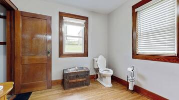 Une salle de bain avec un plancher en bois, une porte en bois, une fenêtre avec des stores, des toilettes et un porte-serviettes en métal.