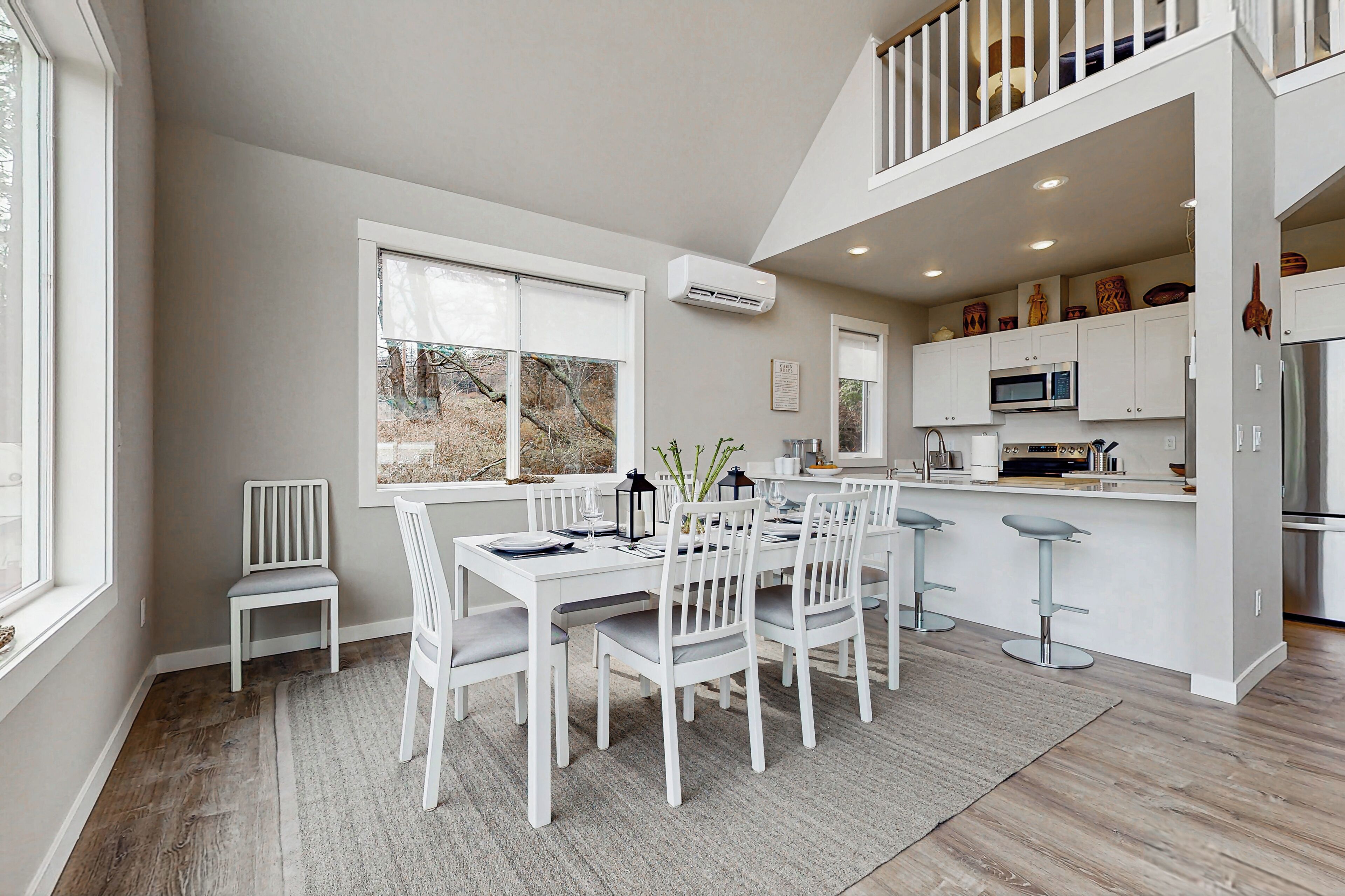 A modern kitchen with white cabinetry, a dining area with a white table and chairs, and a view of the outdoors through large windows.