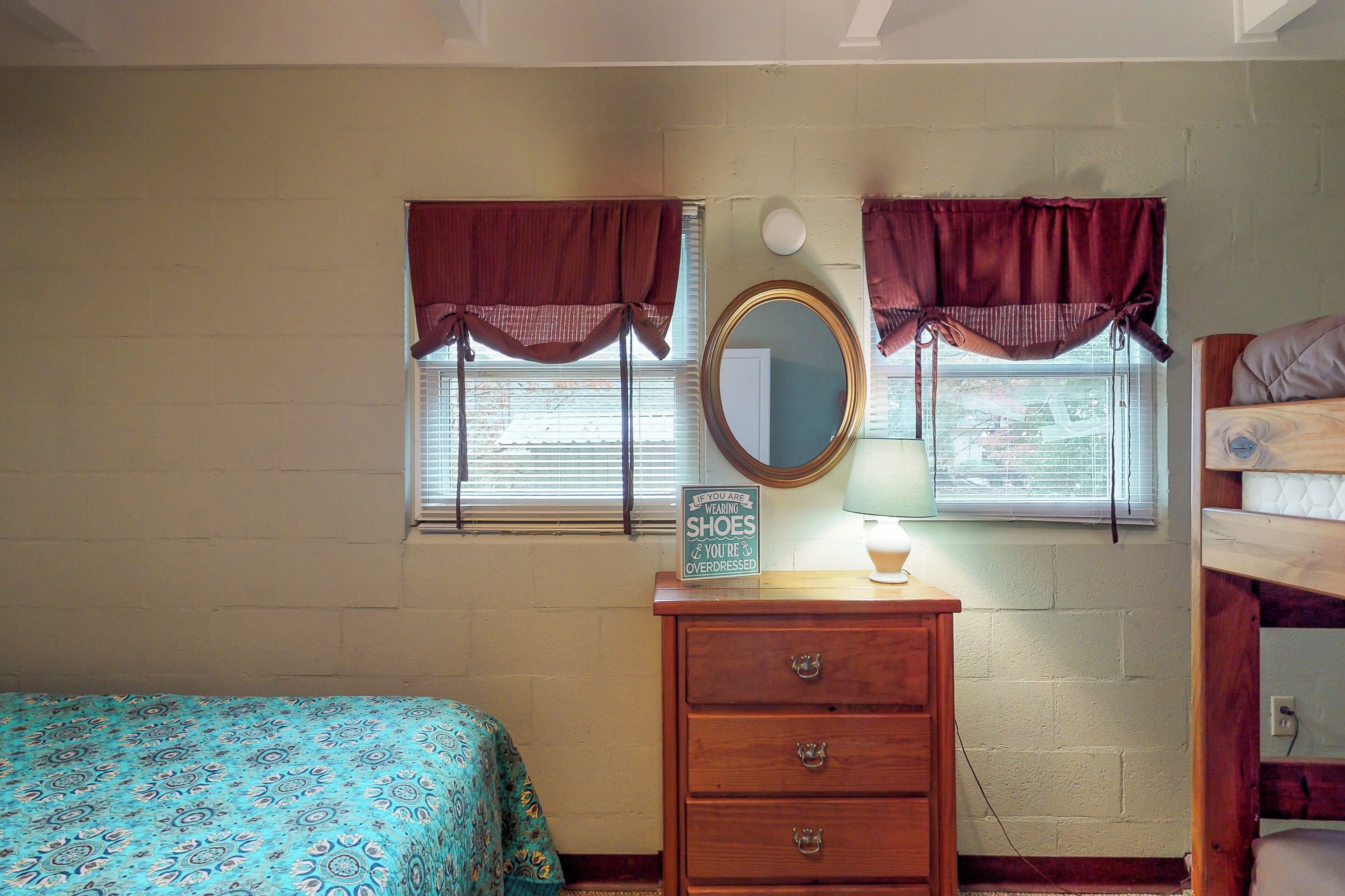 A bedroom with a bed, a dresser, a mirror, and two windows with curtains.