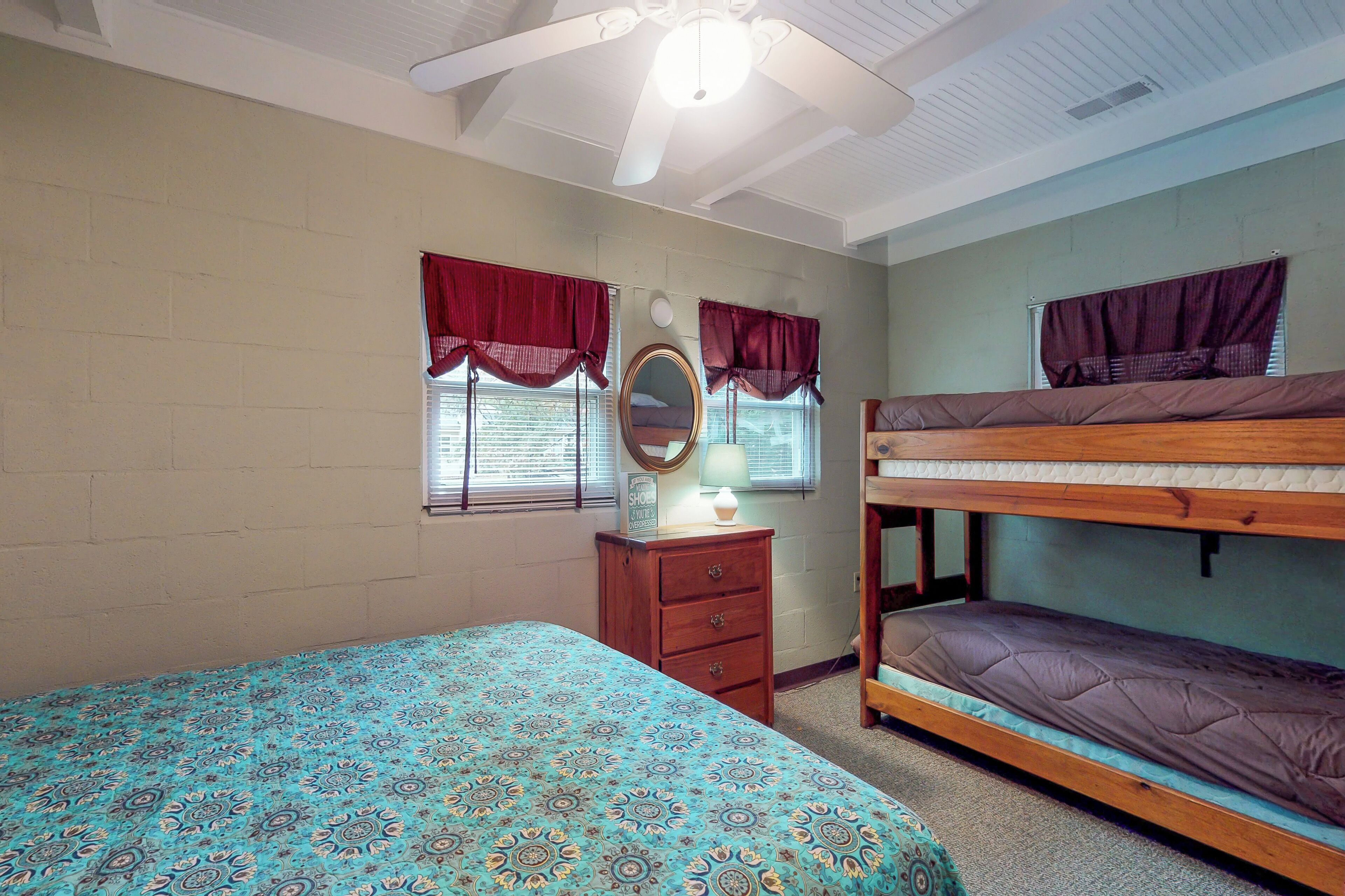 A bedroom with a bunk bed, a single bed with patterned bedding, a dresser, a mirror, and two windows with red valances.