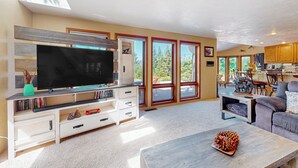 A spacious living room with a large flat-screen TV, a coffee table with a pinecone decoration, and a view of the outdoors through large windows. - 100 Yards to Foster Lake! Family Home w/ Hot Tub (Foster)
