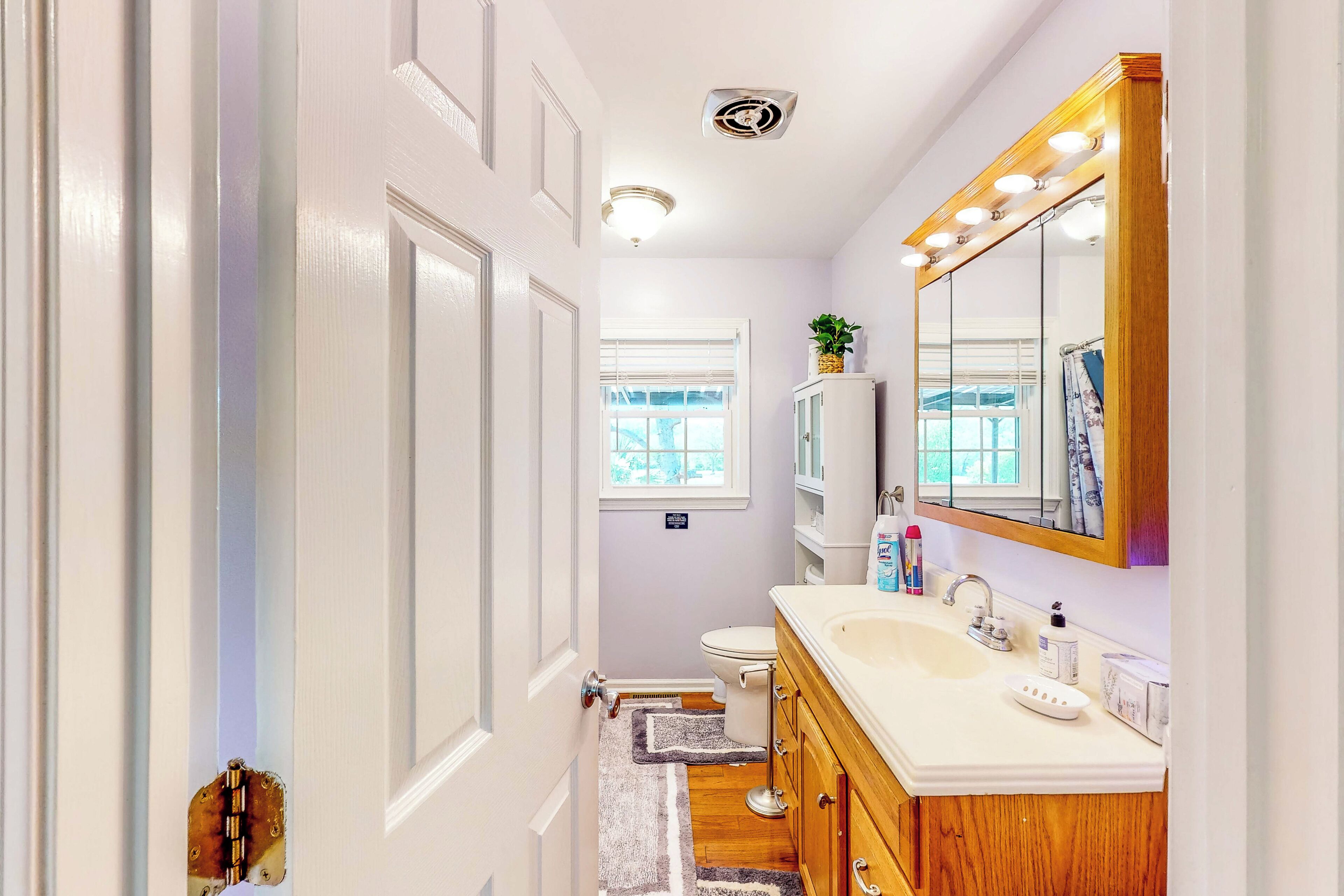 A bathroom with a white vanity, a large mirror with lights, a toilet, and a window.