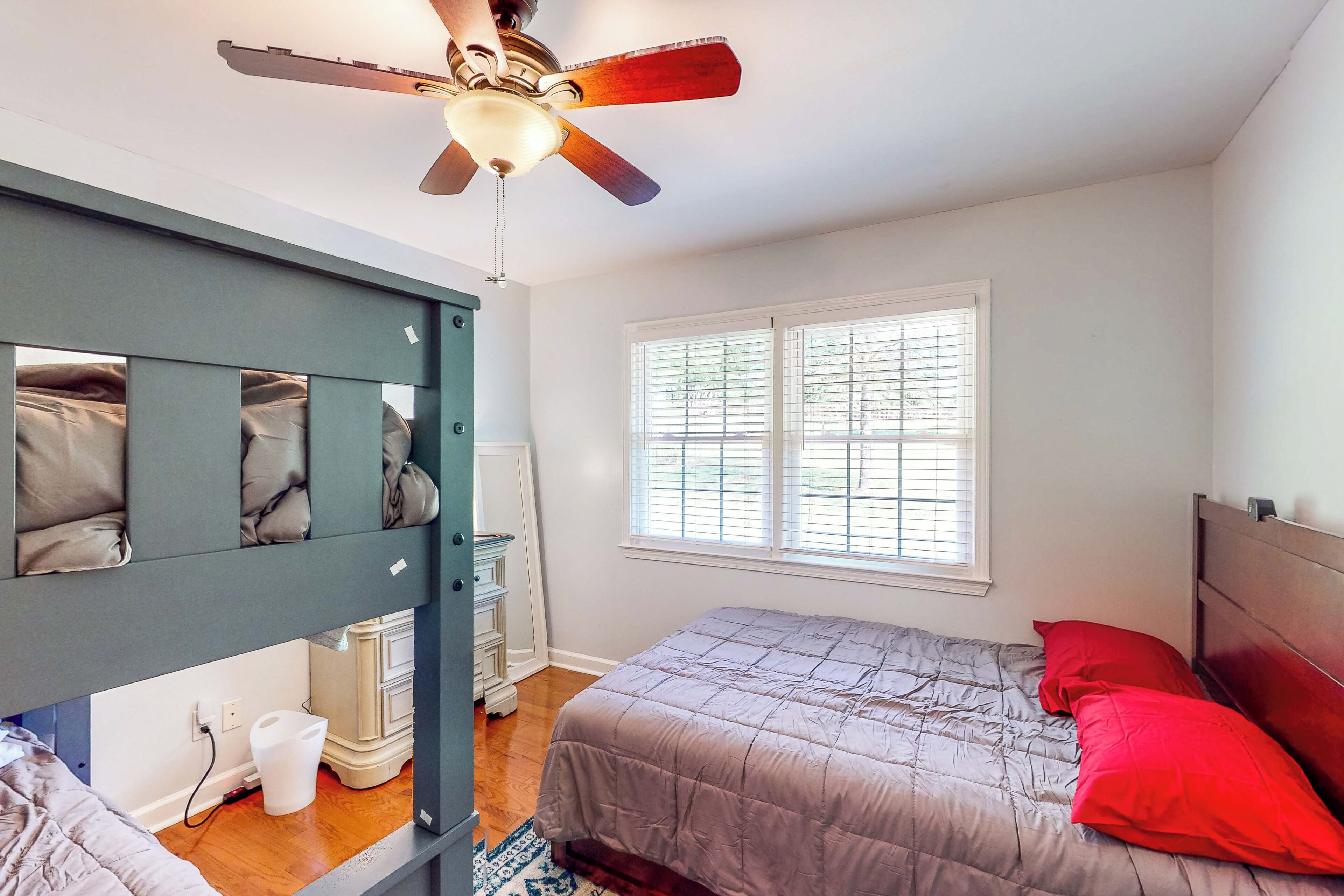 A bedroom with a bunk bed, a single bed with a red pillow, a ceiling fan, and a window with blinds.