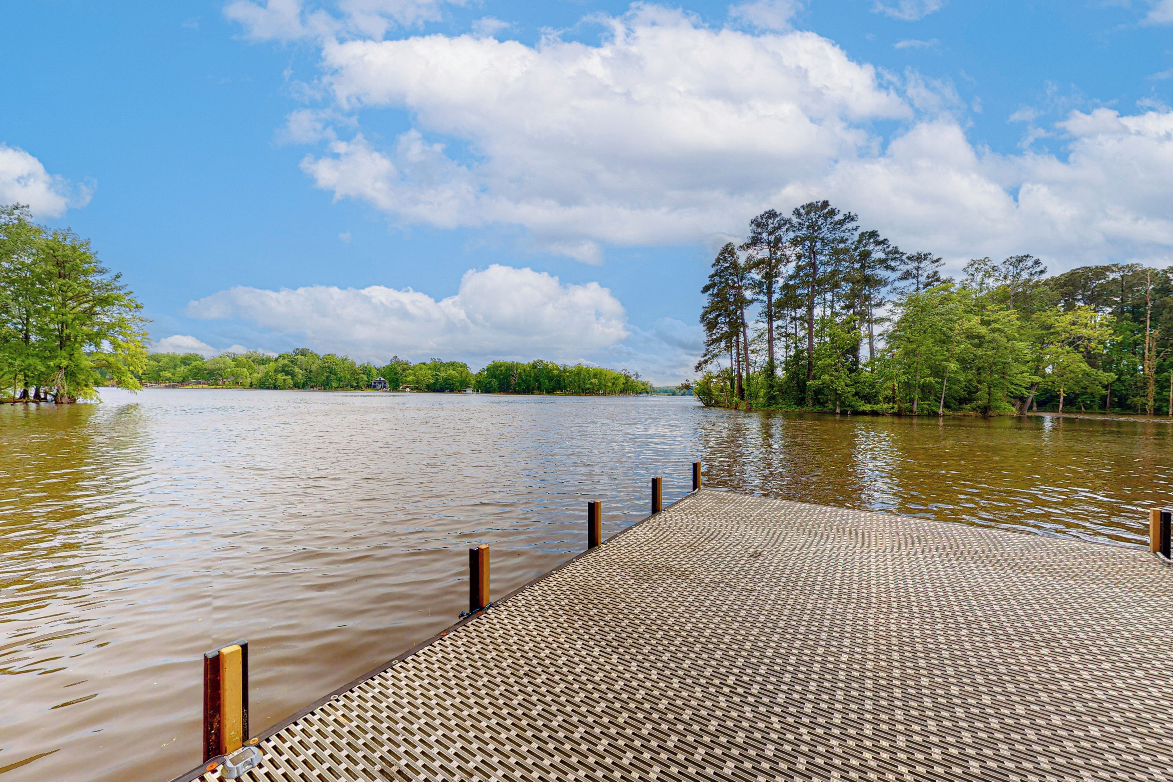 A wooden dock extending into a calm body of water, surrounded by lush greenery and tall trees under a partly cloudy sky.