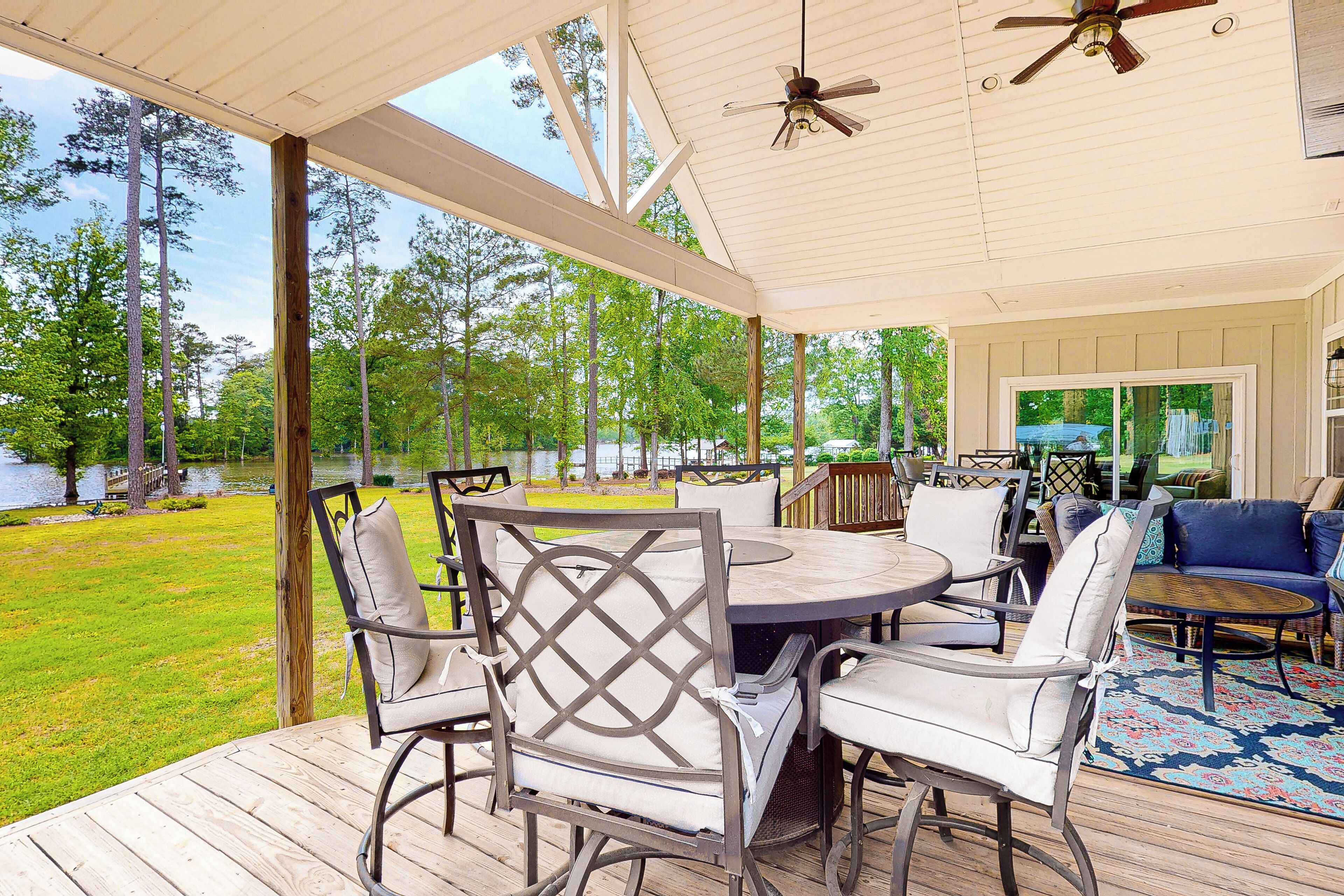 A covered outdoor patio with a wooden deck, a round table with chairs, and a view of a lake and trees.