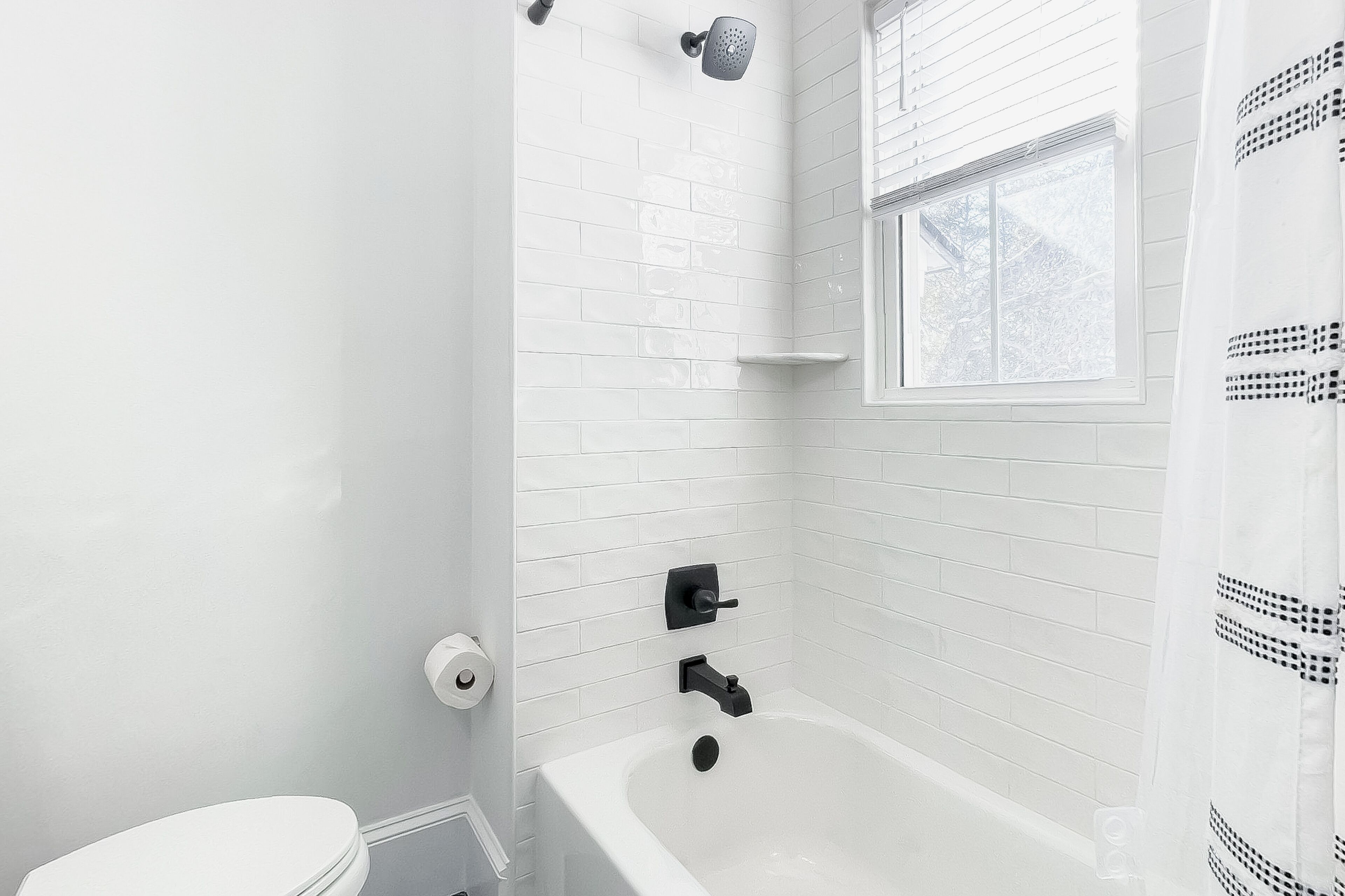 Un baño con paredes cubiertas de azulejos blancos, una bañera blanca, una ventana y un cabezal de ducha.