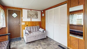 A room with a grey sofa, wooden walls, a white door with a window, and a white closet with louvered doors. - Countryside Couples Retreat! Gateway to Corning (Cameron Mills)