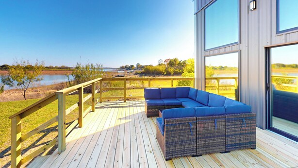 Una terraza moderna al aire libre con un sofá seccional azul y mobiliario de mimbre, con vistas a un cuerpo de agua y una zona verde.