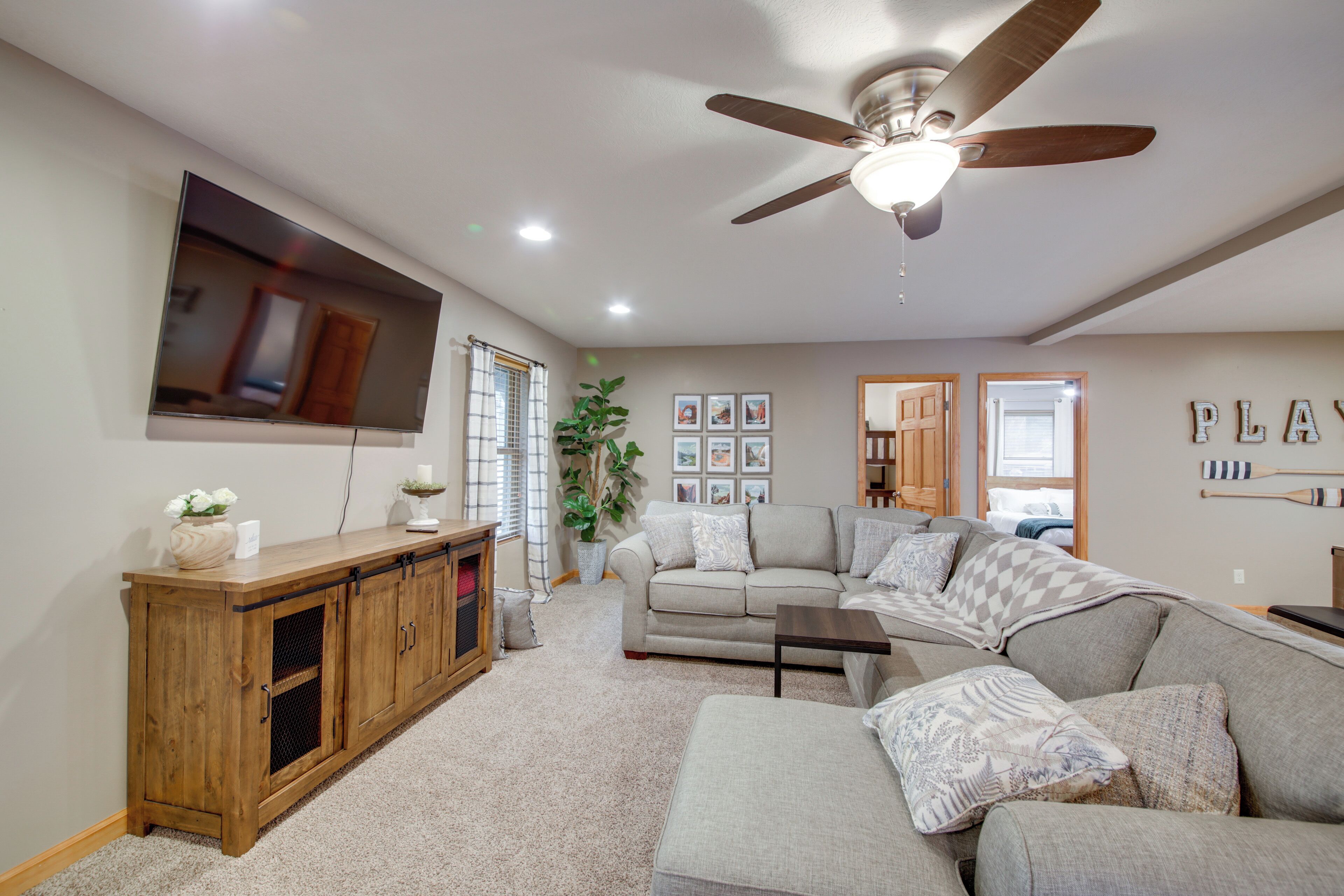 A living room with a large sectional sofa, a wooden cabinet, a ceiling fan, and a flat-screen TV mounted on the wall.