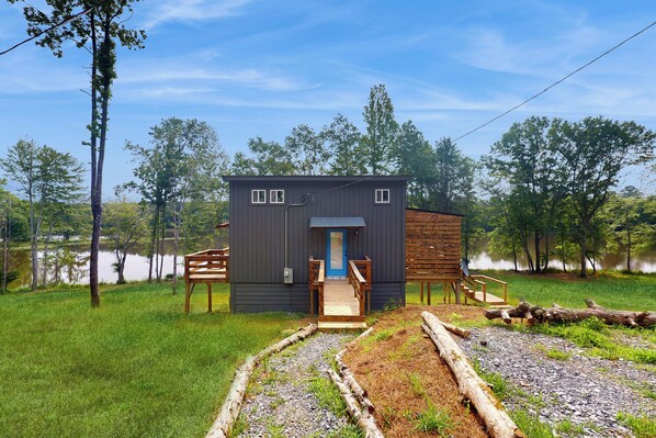 A small cabin with a wooden deck and a blue door, surrounded by trees and a grassy area. - River-view Home w/ Fire Pit & Grill! (Cedar Bluff)