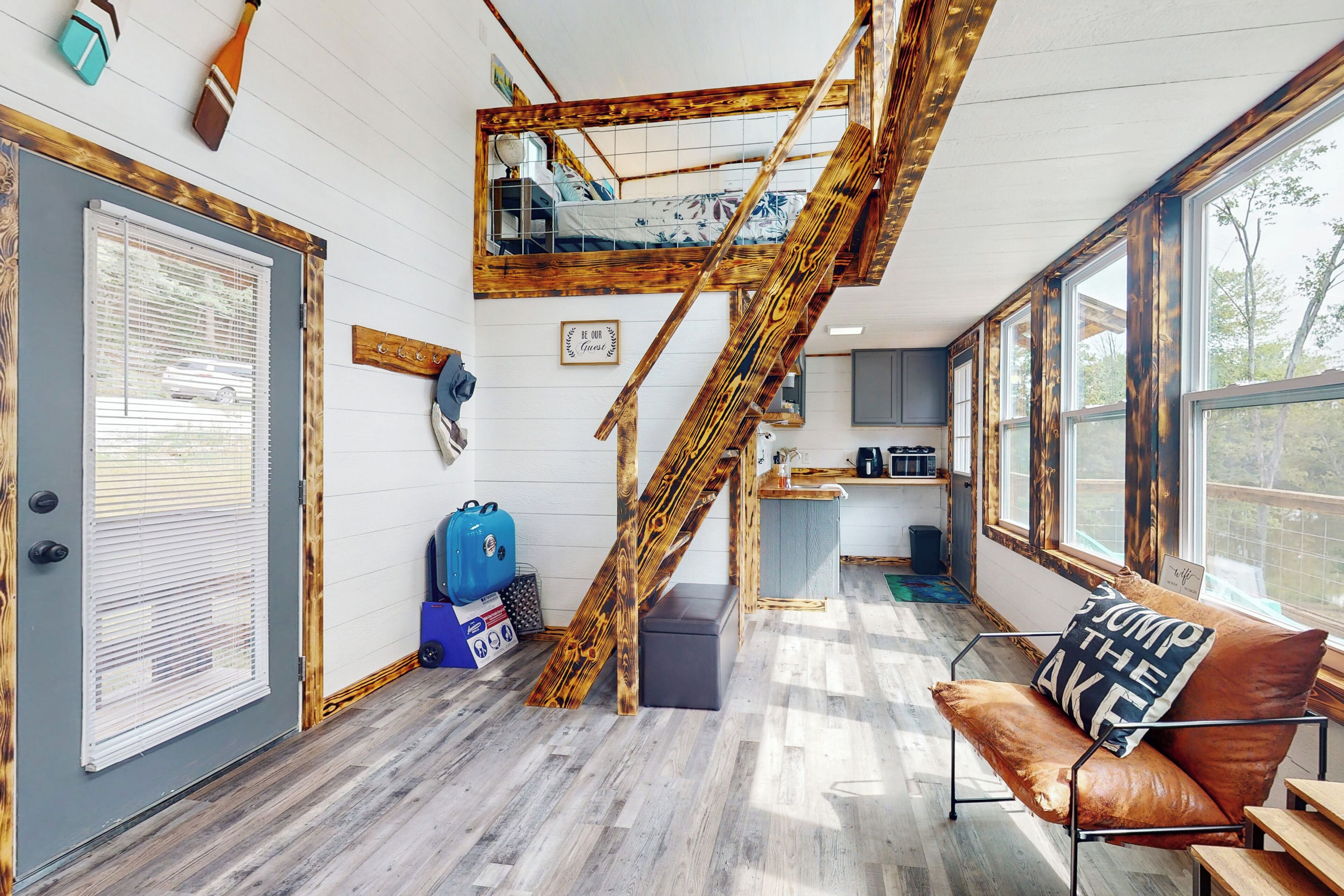 A loft-style room with a wooden staircase, a loft bed, a sofa, and large windows.