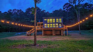 A two-story wooden cabin with a balcony, surrounded by a string of lights and trees. - River-view Home w/ Fire Pit & Grill! (Cedar Bluff)
