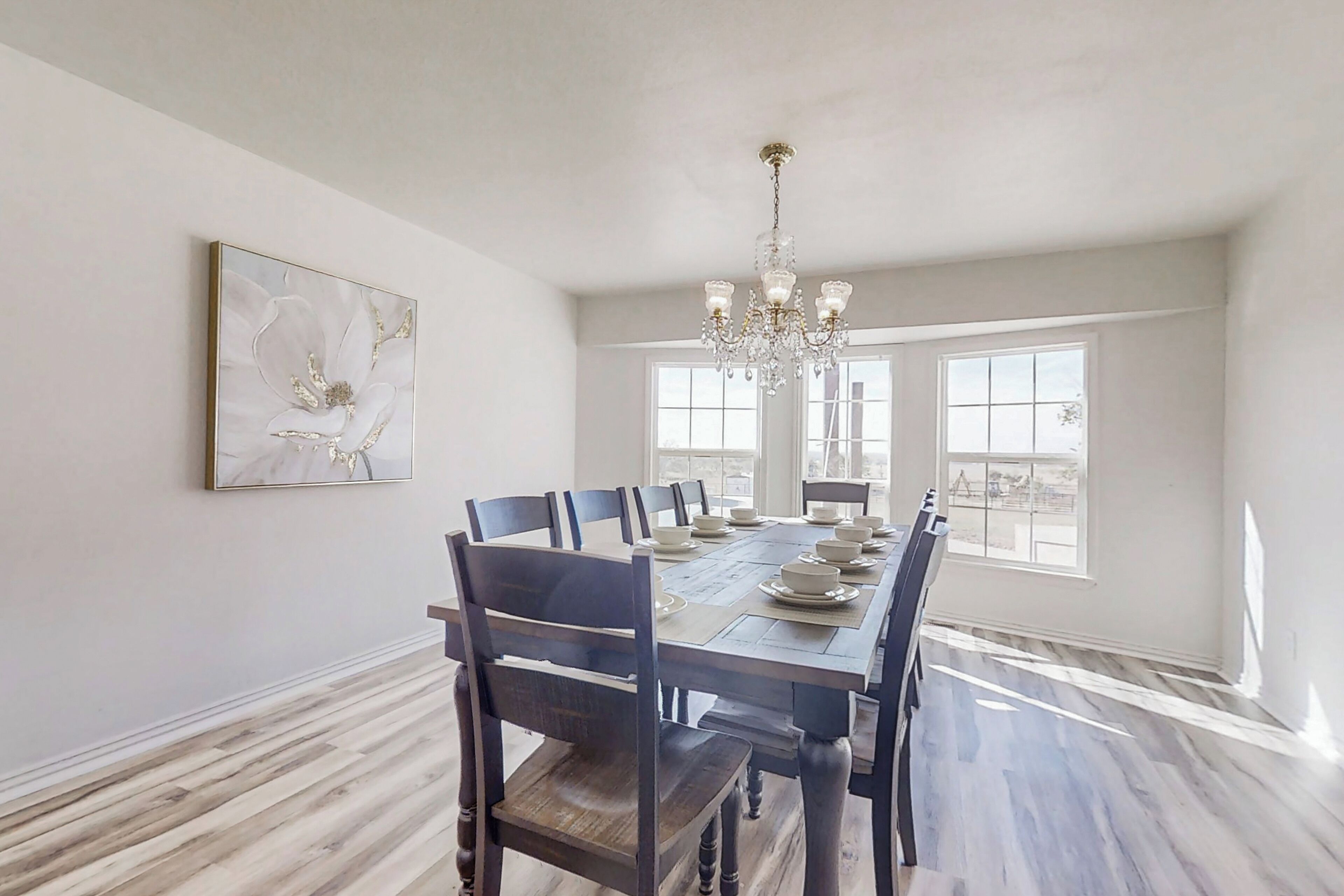 A dining room with a long table set for six, surrounded by chairs, a chandelier, and a large window.