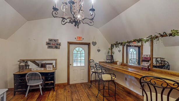 A room with a wooden floor, a desk with drawers, a chair, a chandelier, and a door with an exit sign. - Serene 3BR Farmhouse w/ Deck & Pond (Wilmer)