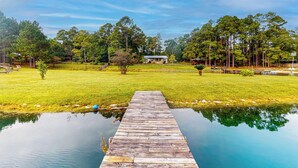 A wooden dock extending into a calm body of water, surrounded by lush greenery and trees. - Serene 3BR Farmhouse w/ Deck & Pond (Wilmer)