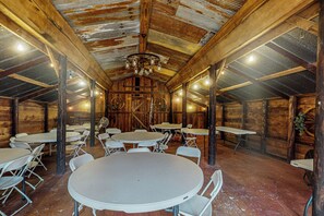 Une salle de style rustique, aménagée comme une grange, avec de nombreuses tables et chaises, des poutres en bois et un ventilateur de plafond.