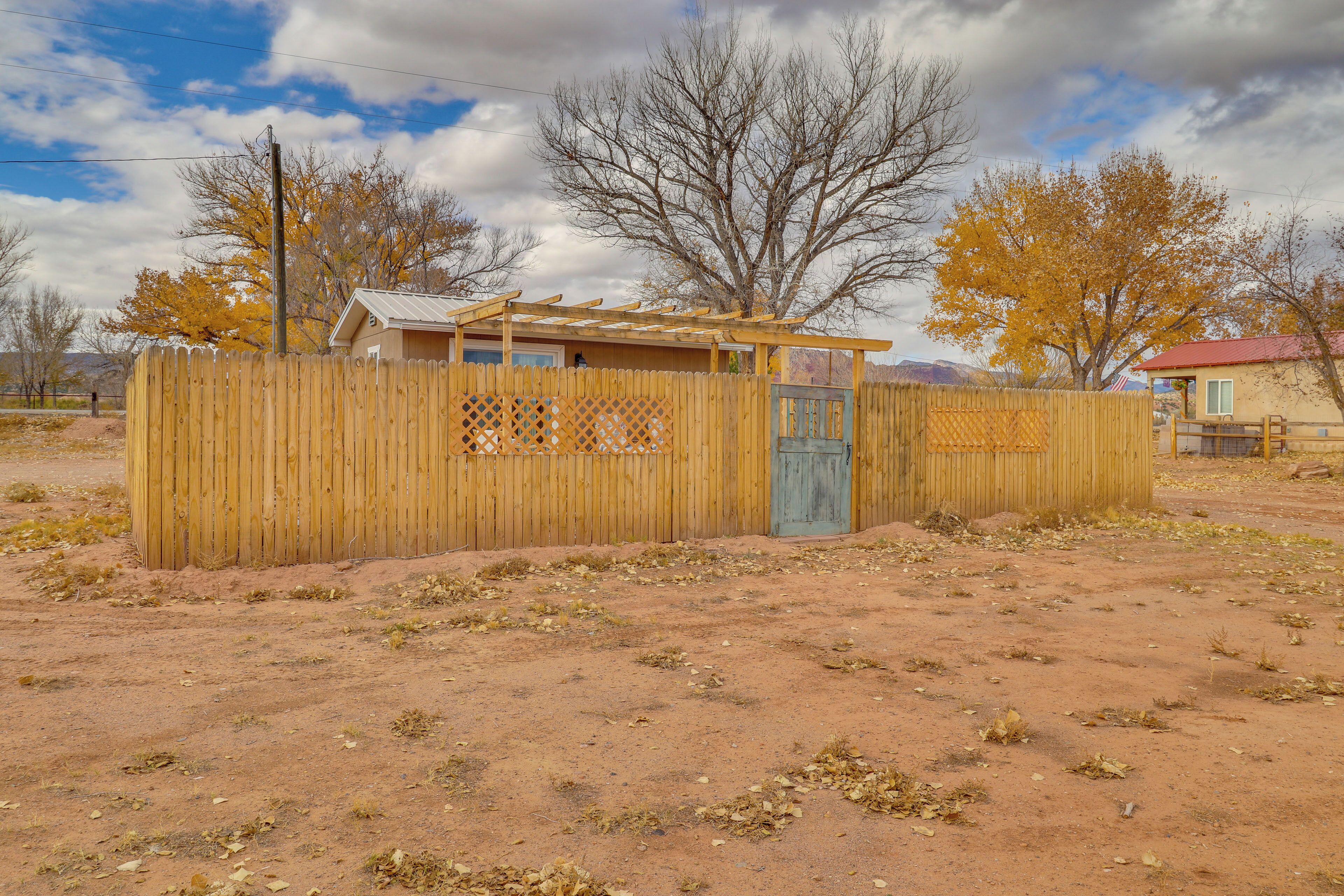 A fenced area with a wooden structure, possibly a shed or small building, in a desert-like environment with sparse vegetation and leafless trees.