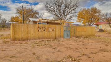 A fenced area with a wooden structure, possibly a shed or small building, in a desert-like environment with sparse vegetation and leafless trees.