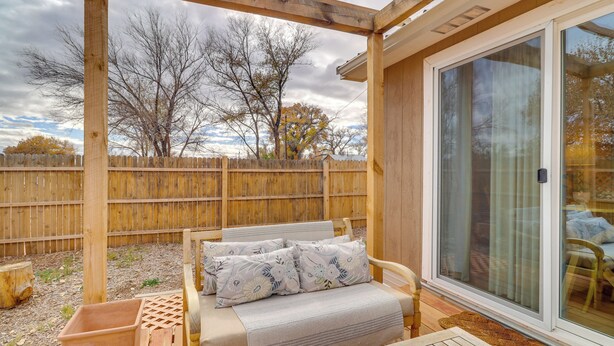 Un patio couvert avec une terrasse en bois, un coin salon confortable avec un banc et des coussins, et une porte-fenêtre coulissante donnant sur un espace extérieur aménagé pour se détendre.