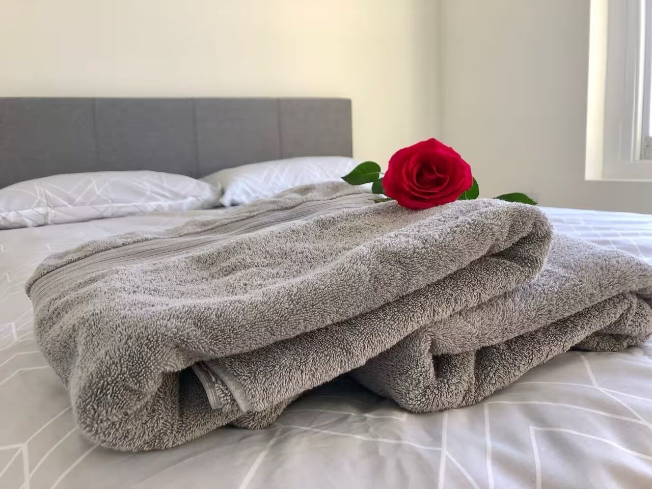 Ein ordentlich bezogenes Bett mit grauer Decke und einer roten Rose darauf.