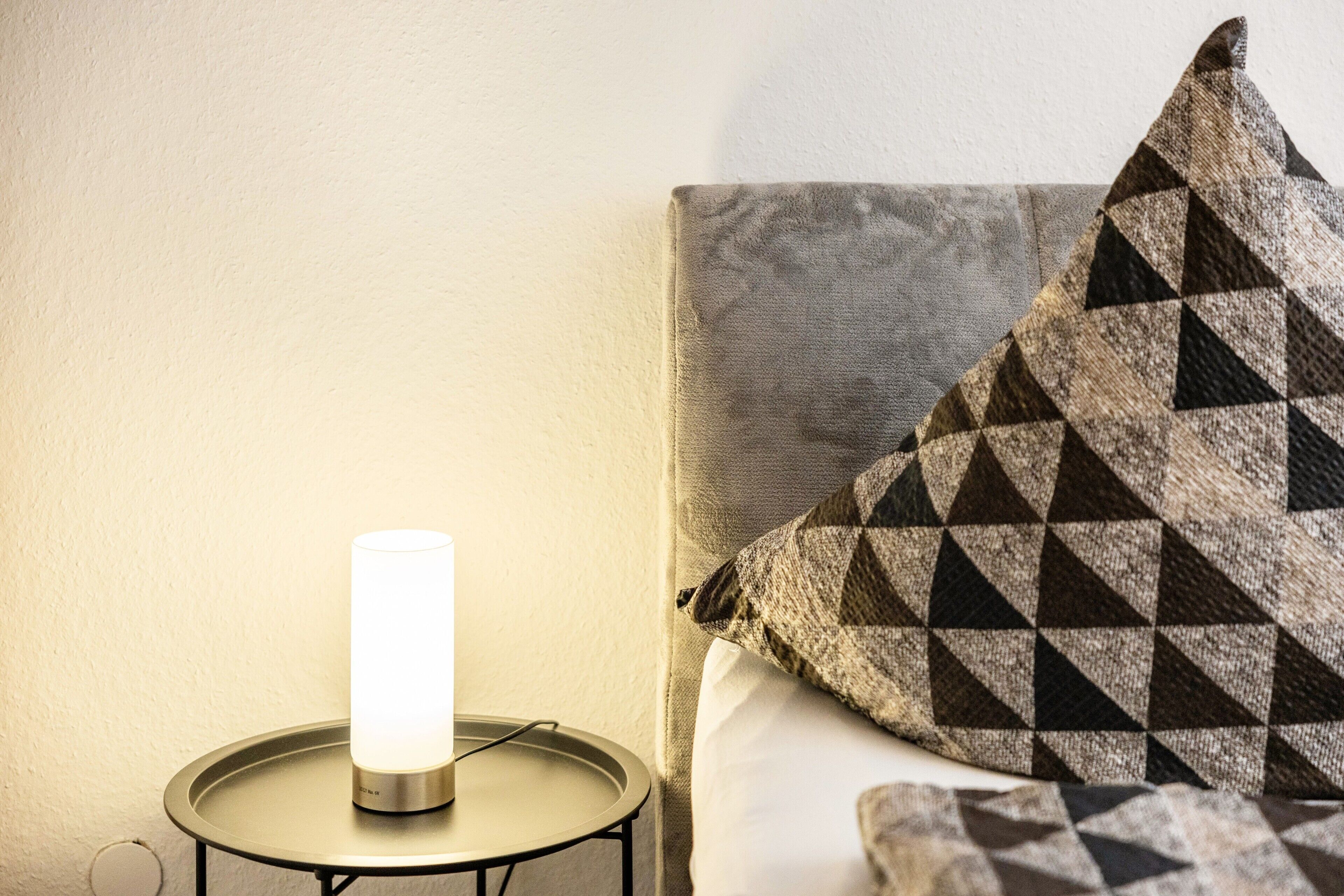 Un comodino con una lampada, un cuscino con motivo geometrico e una parete liscia.