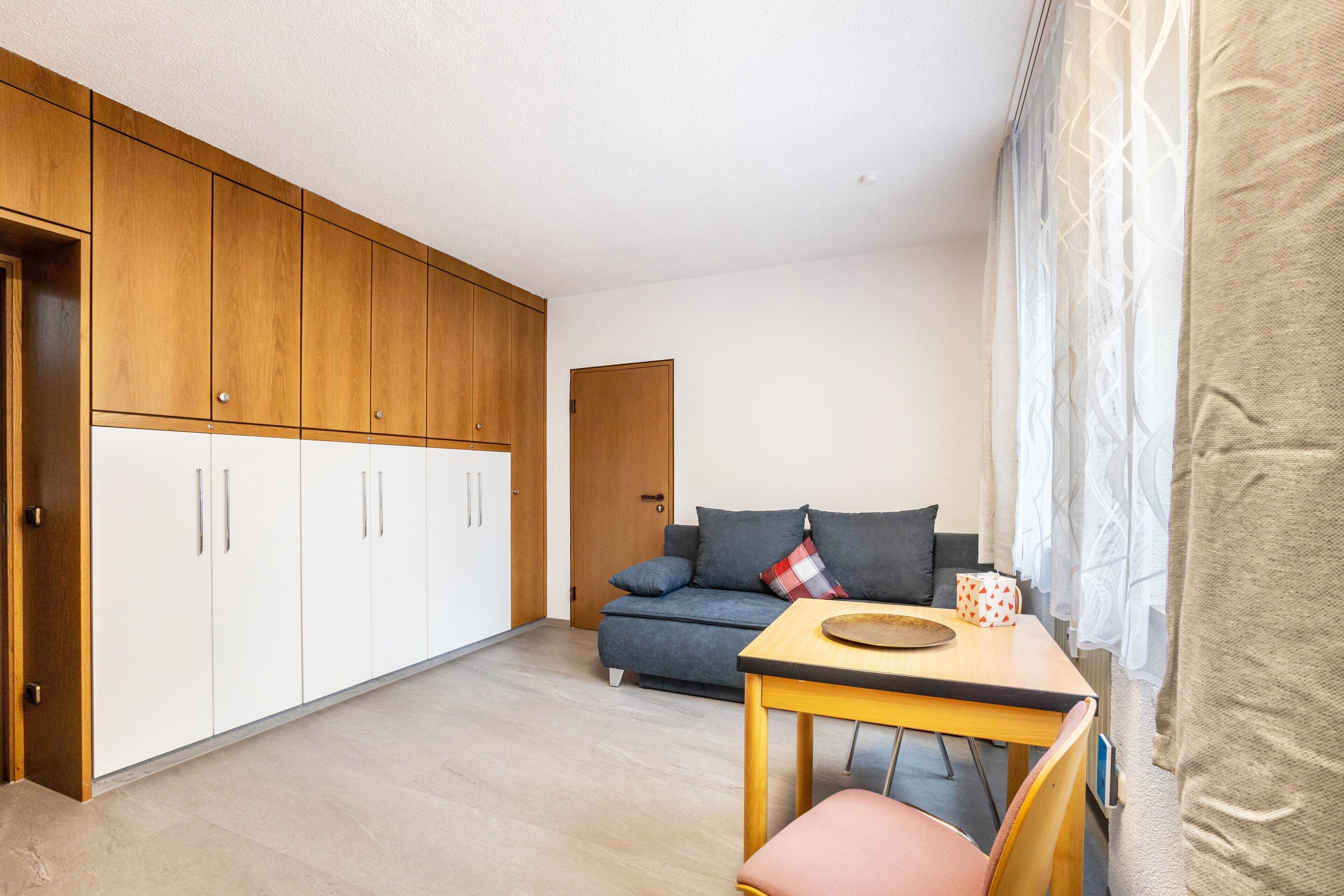 Quarto compacto com cozinha americana, um sofá, uma mesa com cadeira e uma porta que dá para outro espaço.