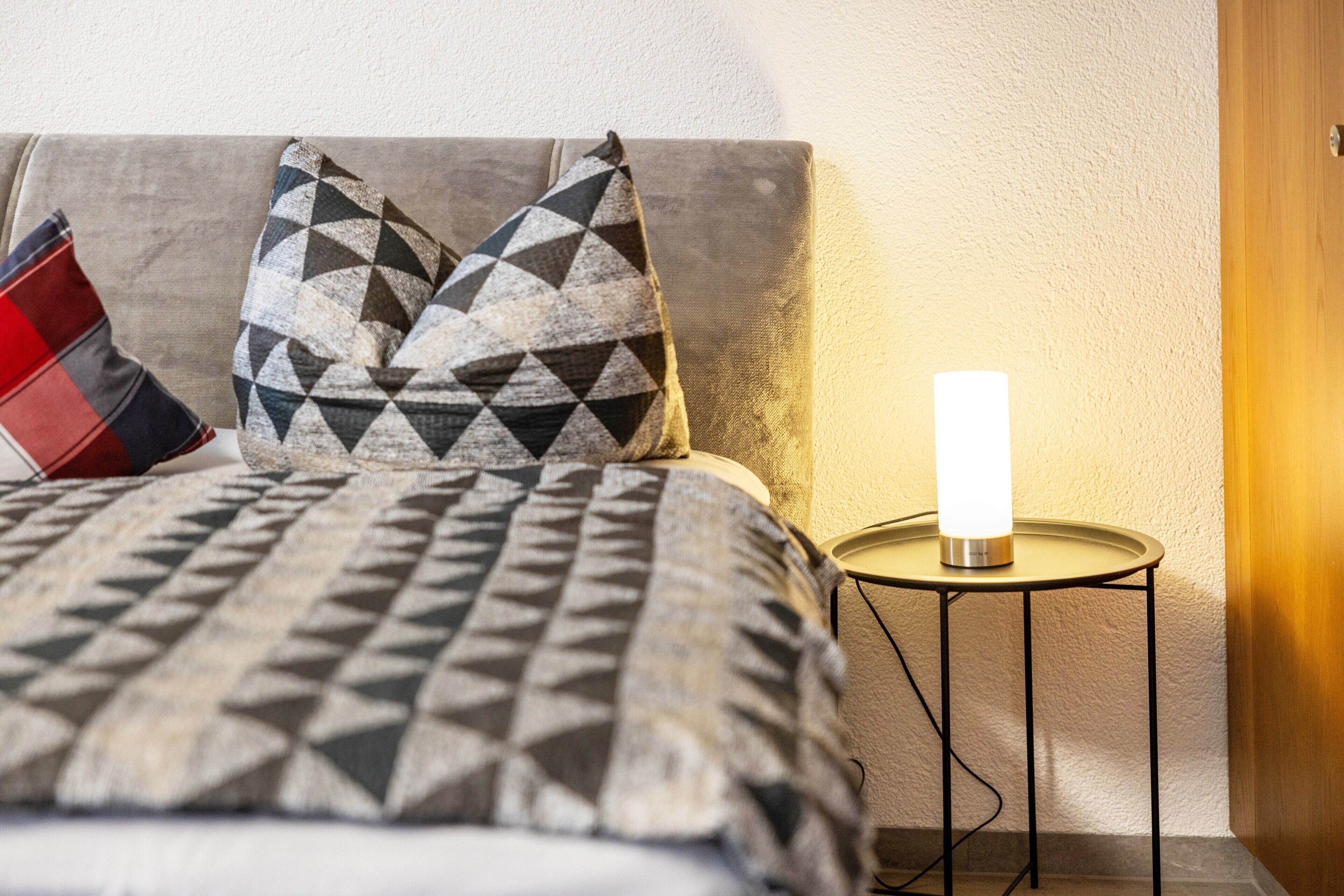 Una cama con ropa de cama estampada, un almohajar rojo y negro, y una lámpara de noche sobre una mesita.