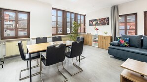 Una sala de estar moderna con un sofá, una mesa de comedor con sillas, una planta en maceta y ventanas grandes.