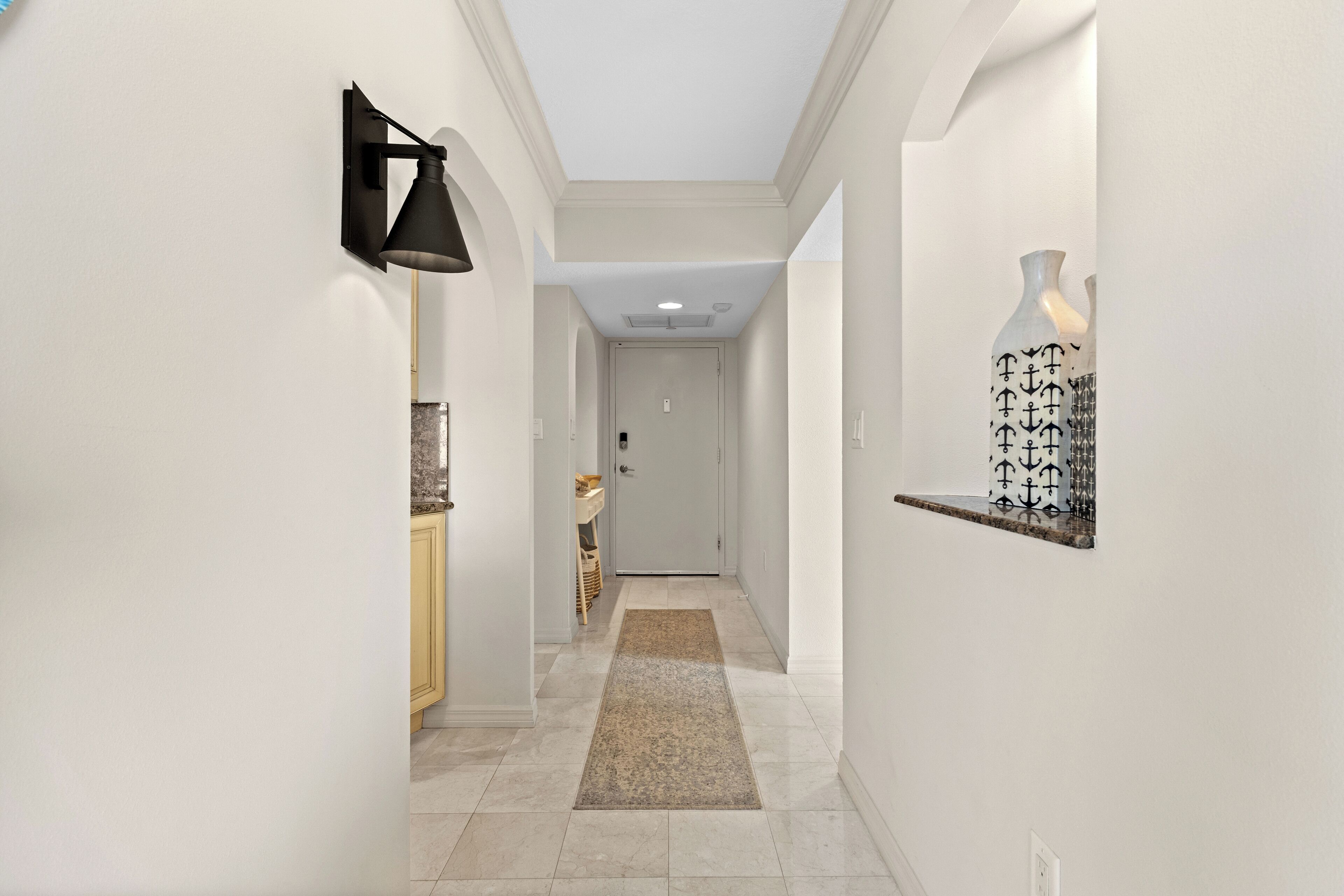 A hallway with a black wall-mounted lamp, a beige rug, and a shelf with decorative bottles.