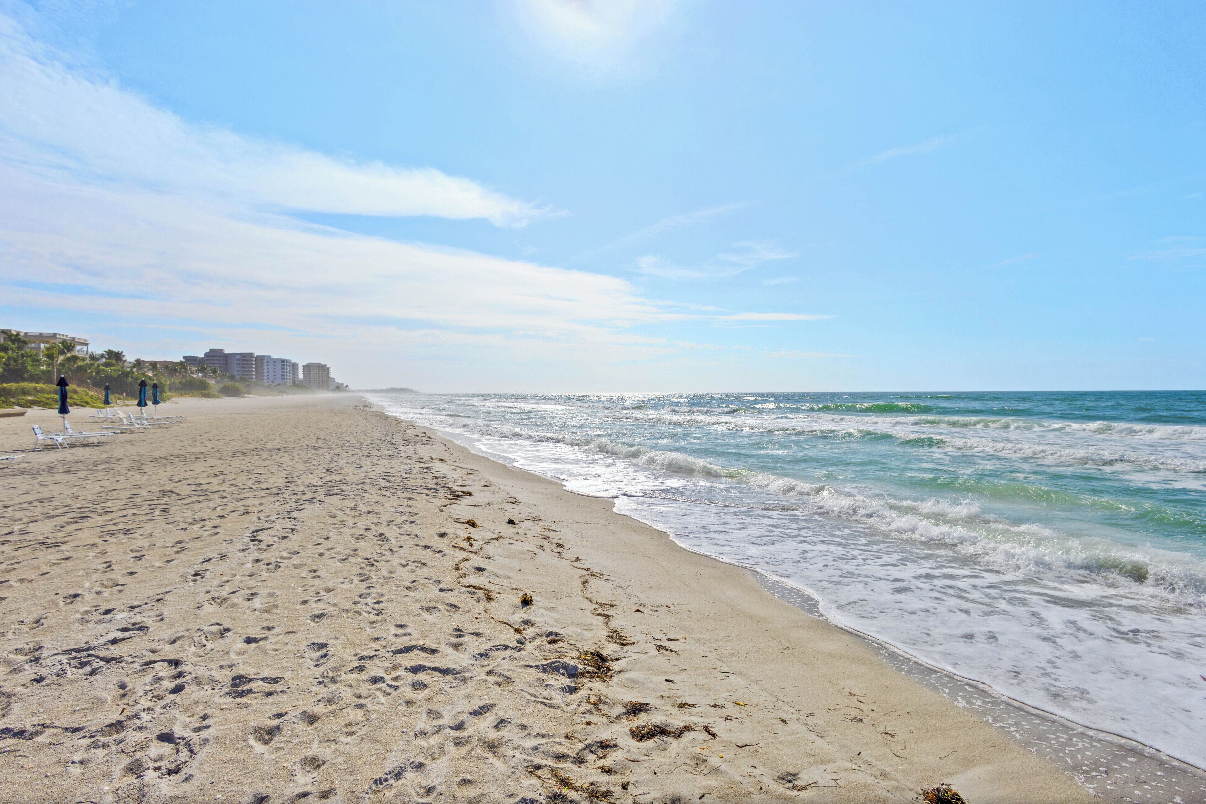 Une plage de sable avec des empreintes de pas, une mer calme et des bâtiments en arrière-plan.
