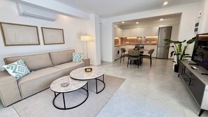 A modern apartment with a living room featuring a sofa and two round tables, a kitchen with a dining area, and a small balcony with a potted plant. - Kantara Apartment w Pool by TrulyCyprus (Limassol)