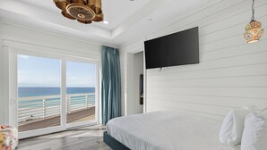 Quarto com uma cama grande, TV fixada na parede e porta de vidro deslizante para uma varanda com vista para o oceano.