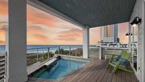 Um deck de madeira com piscina, uma cadeira verde e vista para o oceano ao pôr do sol.