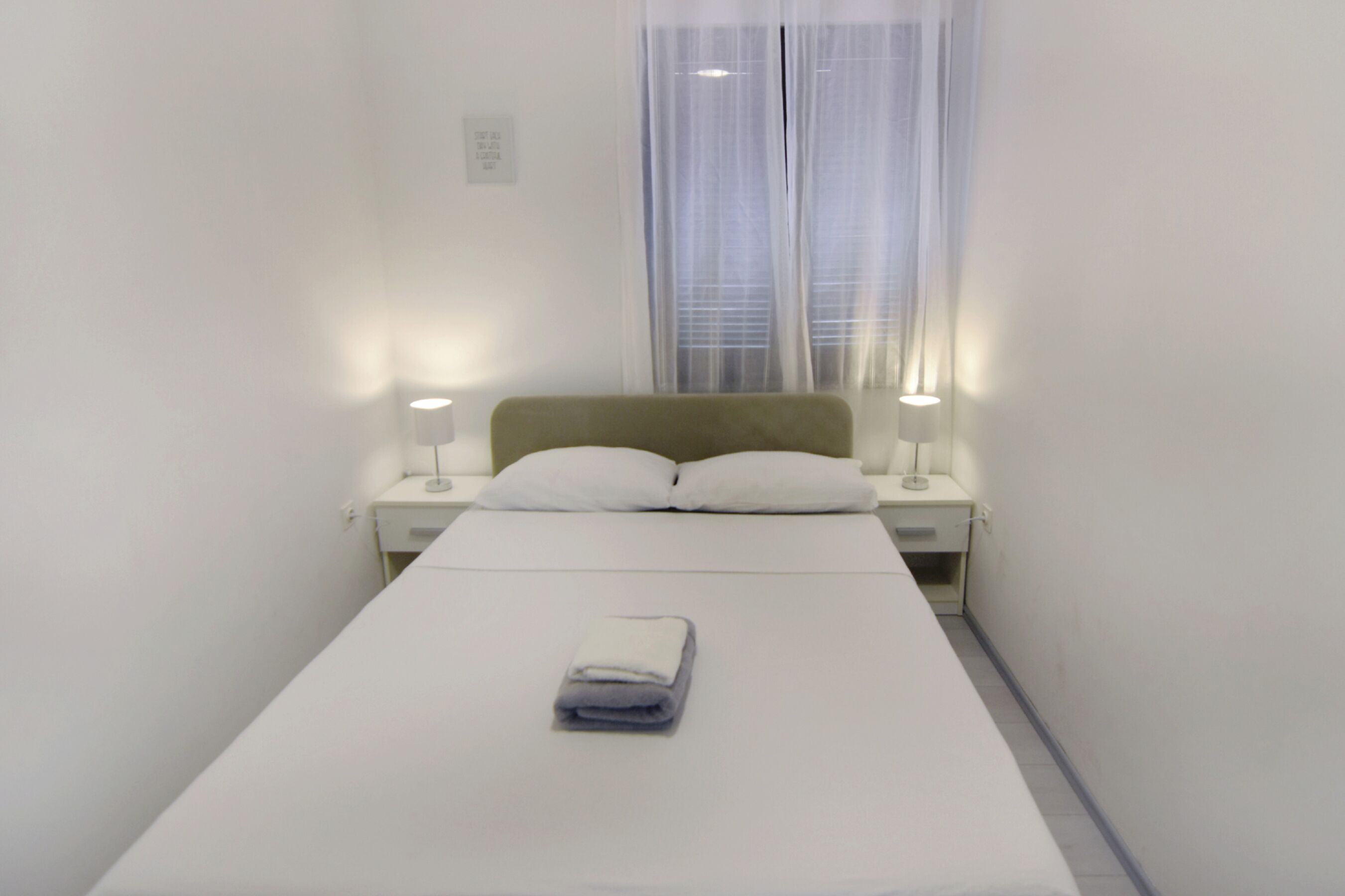 A single bed with a neatly folded towel, two bedside lamps, and a window with sheer curtains.