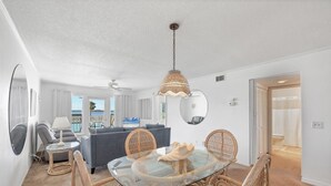 Een eetgedeelte met een tafel met glazen blad, omringd door rieten stoelen, een plafondventilator en een hangende rieten lamp.